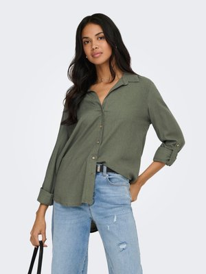 Mujer con pelo largo y oscuro vistiendo una camisa verde oliva con botones y vaqueros azules claros rasgados, sosteniendo un bolso negro con correa con una mano.