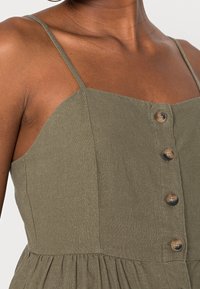 Robe en lin vert olive avec des bretelles fines, présentant un décolleté droit et six boutons sur le devant. Tissu doux et texturé avec des plis.