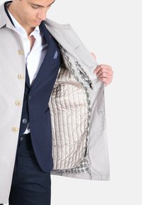 Cappotto beige chiaro con grandi bottoni, caratterizzato da una fodera interna trapuntata color crema e dettagli a quadri. La modella mostra l'interno con una mano.