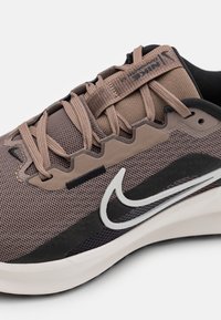 Scarpe Nike con tomaia in mesh marrone, sovrapposizioni testurizzate, dettagli scuri e un prominente logo swoosh bianco. Presentano lacci color crema e una suola imbottita.