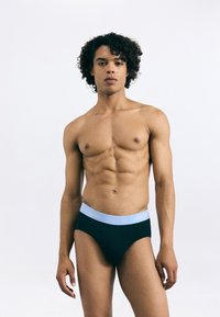 Fekete pamut bugyi világoskék elasztikus derékpánttal, amelyen a "Calvin Klein" logó látható. A modell magabiztosan pózol mezítelen torsóval.