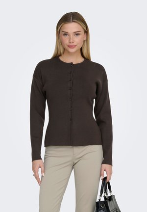 Ženska z rjavim gumbom na rjavi jopi in bež hlačami, ki drži črno torbico s kratkimi ročaji na belem ozadju.