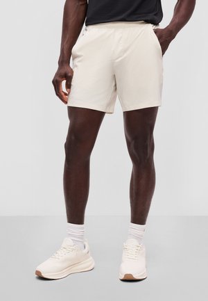 Mann trägt hellbeige Shorts, weiße Socken und hellbeige Sneakers und steht vor einem einfarbigen grauen Hintergrund.