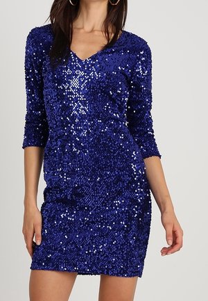 Kobieta ubrana w dopasowaną sukienkę z królewskiego niebieskiego cekinowego materiału, z rękawami do łokcia i dekoltem w kształcie litery V, stojąca na tle jednolitego jasnego tła.