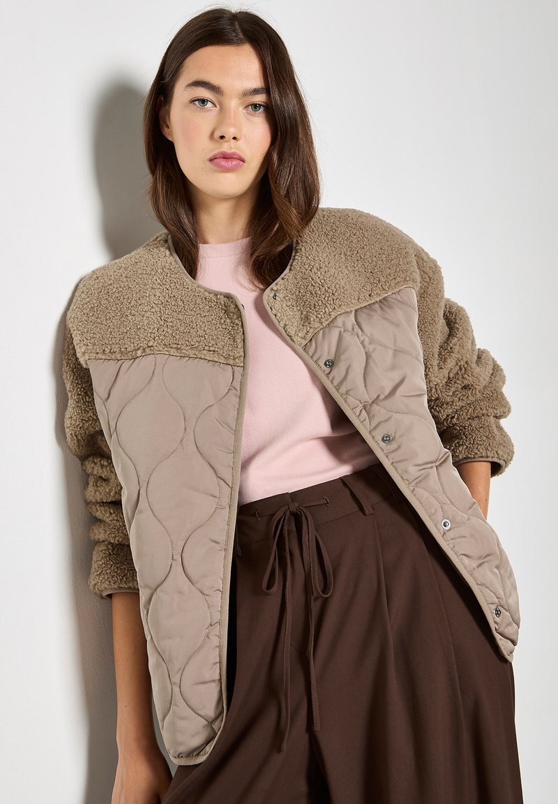 Femme aux cheveux bruns portant une veste matelassée et en polaire beige sur un haut rose et un pantalon marron taille haute, debout contre un mur blanc.