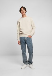 Sudadera beige de manga larga y cuello redondo, combinada con unos vaqueros azul claro. Detalle destacado del logo en el pecho. Zapatillas blancas.