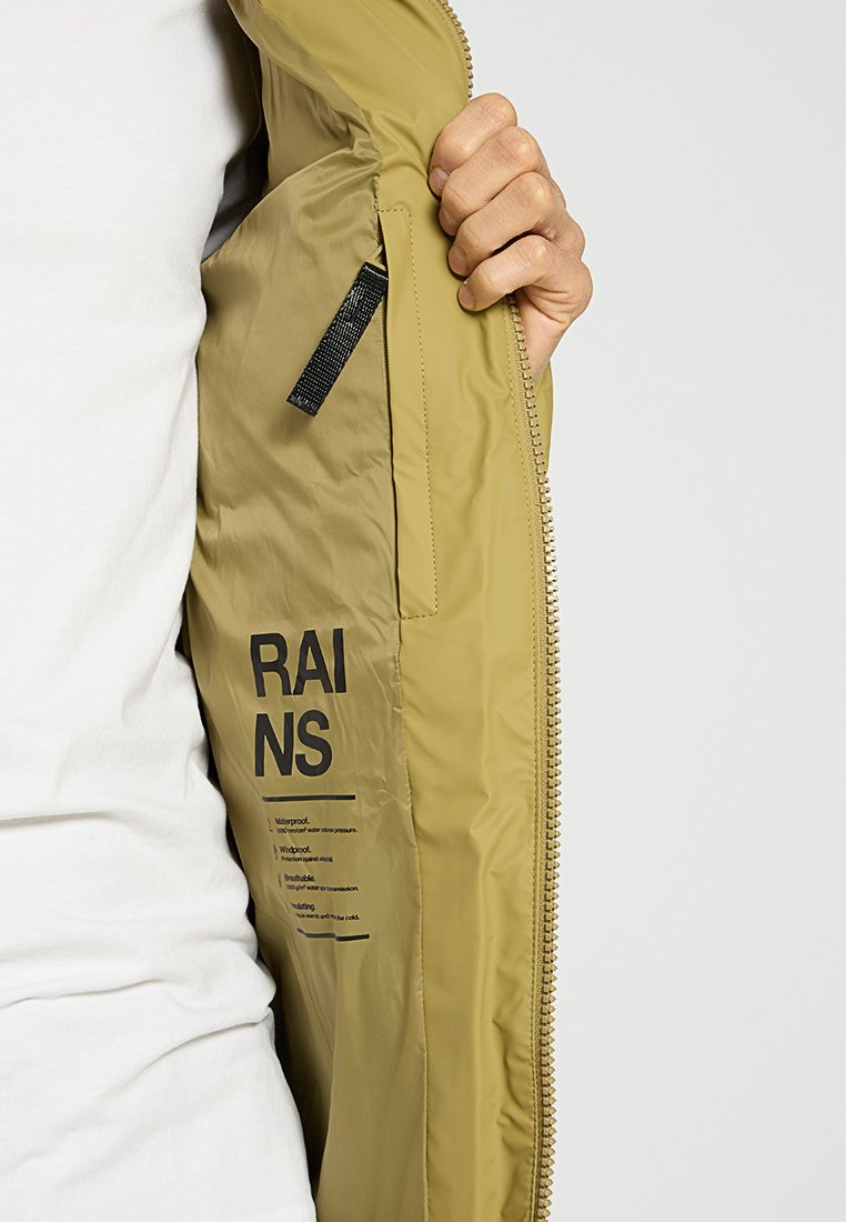 Hand die een beige regenjas opent en het binnenlabel toont met het "RAINS" logo en de tekst waterproof, winddicht, ademend.