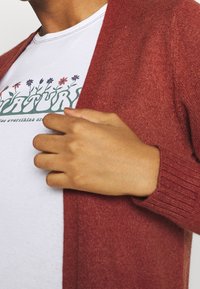 Maroon cardigan med ribbat mönster, öppen front och långa ärmar. Bärs över en vit skjorta med ett blommigt grafiskt mönster.