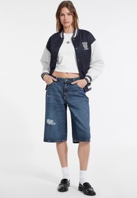 Varsityjack in marineblauw met grijze mouwen, cropped wit topje, versleten denim bermudashorts, witte sokken en zwarte loafers.