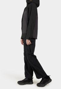 Veste noire avec panneaux gris, poches zippées et capuche. Pantalon cargo noir avec poches, porté avec des baskets noires avec des détails en mesh.