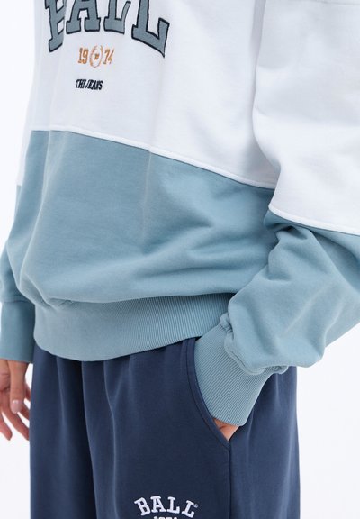 Light blue and white color-blocked sweatshirt with ribbed cuffs, featuring “BALL 1974” in navy and gold embroidery, paired with navy sweatpants.