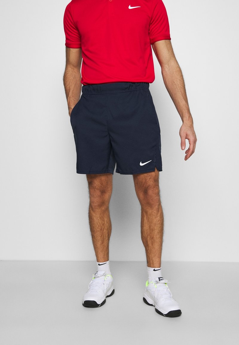 Pantalones cortos atléticos de color azul marino con un diseño sólido, cinta elástica en la cintura, bolsillos laterales y un logo de Nike blanco en la parte inferior izquierda. Combinados con una camiseta polo roja.