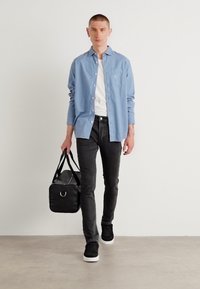 Camisa de botón azul claro sobre una camiseta blanca, jeans negros y zapatillas negras, llevando una bolsa de viaje negra con un acento plateado.