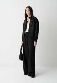 Een vrouw staat zelfverzekerd, gekleed in een zwarte jacket, een wit shirt en losse zwarte broeken, terwijl ze een zwarte schoudertas vasthoudt tegen een minimalistische achtergrond.