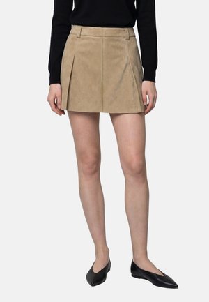 Femme portant une mini-jupe plissée beige, un haut noir à manches longues, et des ballerines noires pointues, debout sur fond blanc.