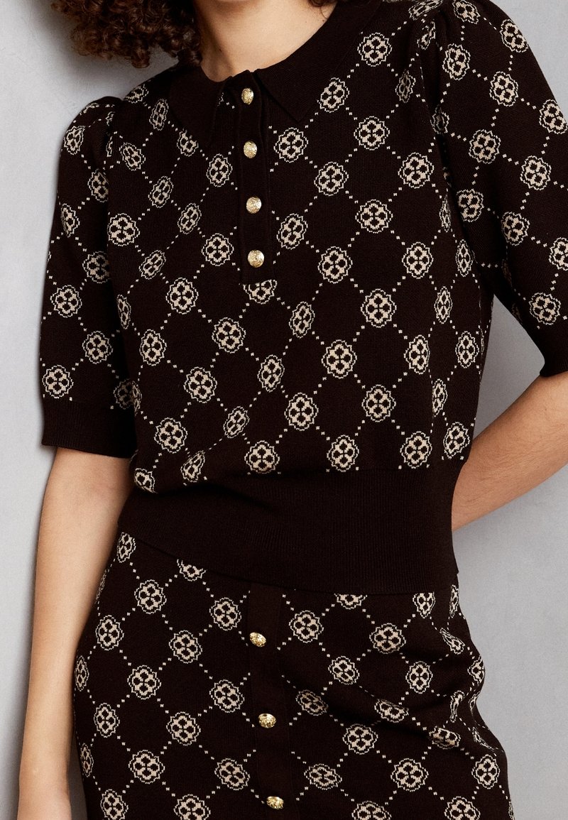 Ensemble haut en tricot marron foncé et jupe avec motif floral losange beige et boutons dorés, porté par une personne aux cheveux bouclés.