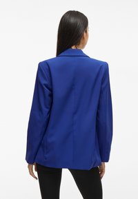 Femme aux longs cheveux foncés portant un blazer bleu roi et un pantalon noir, vue de dos sur un fond blanc.