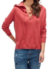 Röd huva tröja med snörning vid kragen och metallöglor, tillverkad av mjukt tyg. Har långa ärmar och en avslappnad passform. Bärs med blå slitna jeans.