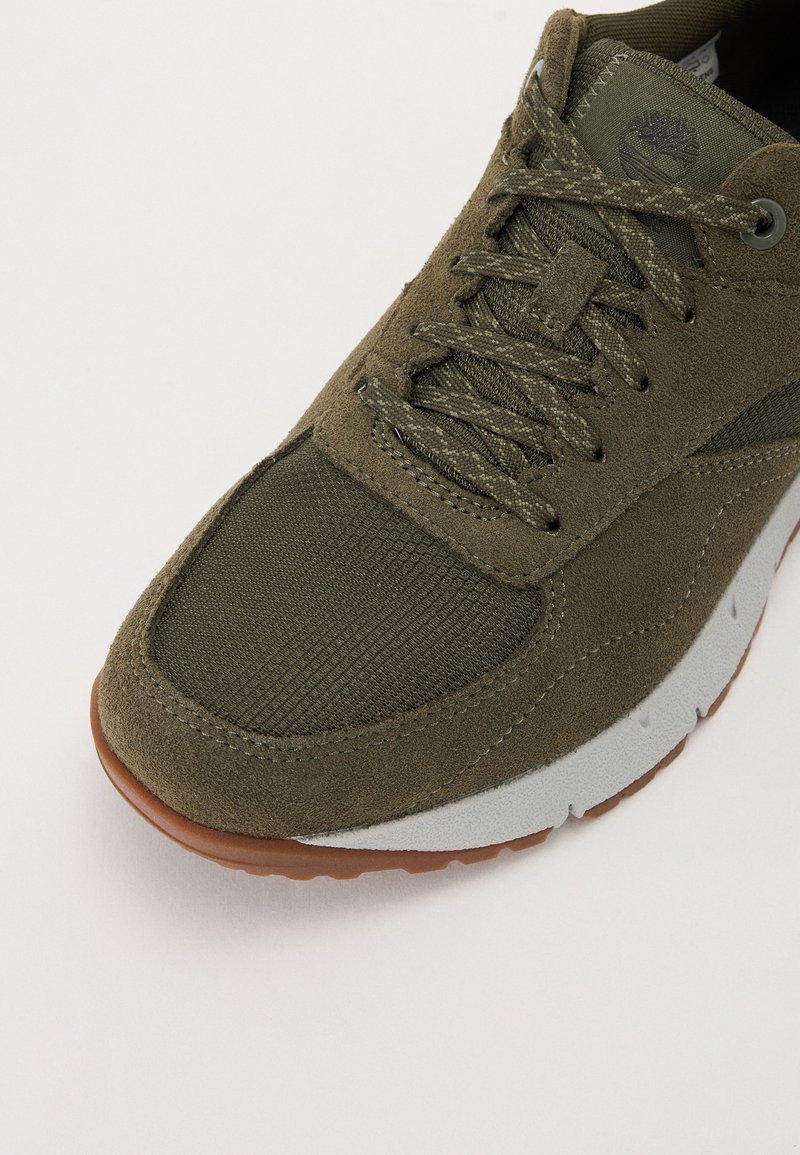 Zapatilla de color verde oliva con texturas de ante y malla, punta redonda, diseño con cordones, entresuela gris y suela de goma marrón.