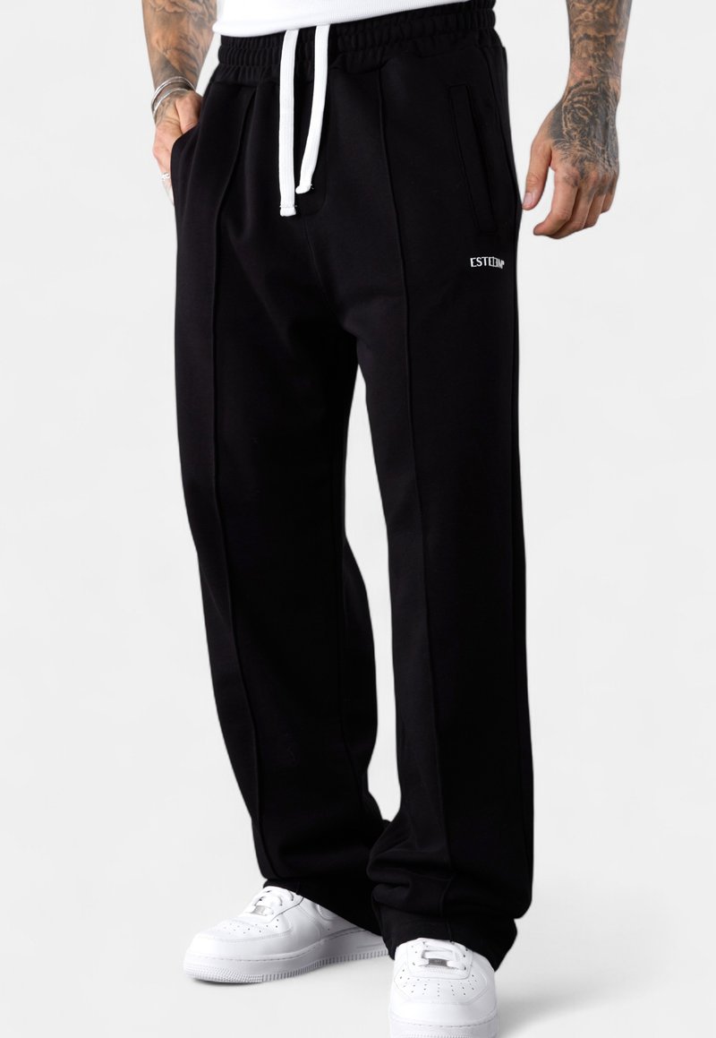 Man wearing black sweatpants with white drawstrings and white sneakers, hands relaxed by sides, showing tattoos on forearms.