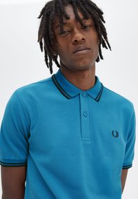 Polo shirt turchese realizzato in un tessuto texturizzato con una chiusura a tre bottoni, colletto a strisce nere e verdi, e un piccolo logo ricamato.