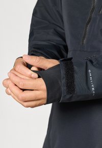 Črna zunanja oblačila z zadrgo spredaj in Velcro prilagodljivimi manšetami. Ima gladko, teksturirano tkivo za fleksibilnost in udobje.