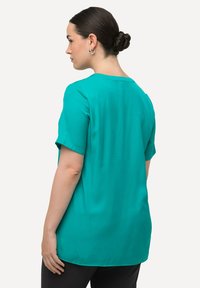 Blusa corta a maniche corte color teal con scollatura rotonda, realizzata in tessuto liscio; presenta una vestibilità rilassata e un delicato dettaglio di piega sul retro.