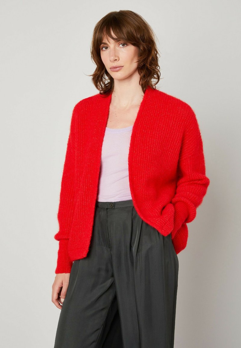 Rdeča pletena jopiči z odprtim prednjim delom, rebrasto teksturo in dolgimi rokavi. Usklajen z svetlo vijoličnim topom in temno plisiranimi hlačami.