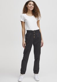 Pantalon noir effilé avec une taille élastique, comportant trois boutons en bois sur le devant. Associe à un T-shirt blanc et des baskets blanches.