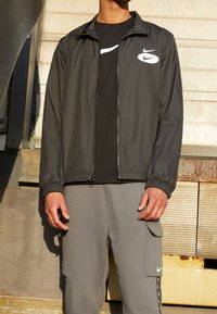 Grijze Nike windbreakerjas met ritssluiting en elastische manchetten, gedragen over een zwart shirt. Gecombineerd met lichtgrijze cargobroek met zakken.