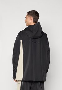 Casaco preto com capuz e um painel lateral bege em contraste, apresentando uma textura suave e um design minimalista. Logótipo exibido nas costas.