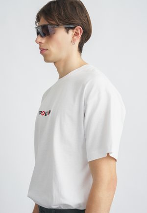 T-shirt blanc en coton à manches courtes et coupe ample, avec un texte brodé en rouge et noir. Modèle portant des lunettes de soleil foncées.