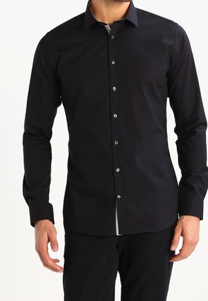 Mann trägt ein eng anliegendes, langärmeliges schwarzes Hemd mit Knöpfen und Kragen sowie schwarze Hose vor schlichter Hintergrundkulisse.