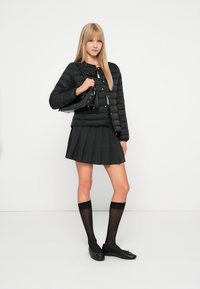 Ung kvinne iført en svart quilted jakke, plisserte svarte skjørt, gjennomsiktige svarte kne-høye strømper, svarte leiligheter, holder en svart håndveske.