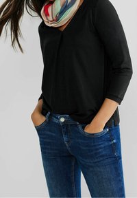 Svart långärmad topp med V-ringning, buren över blå denimjeans. En färgglad mönstrad scarf ger en accent till looken.