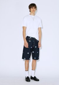 Camiseta blanca, pantalones cortos de mezclilla azul con bordados florales, zapatos negros, calcetines blancos, de pie contra un fondo blanco liso.