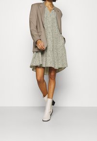 Blazer beige sobre un vestido corto con estampado floral y bajo acampanado. Botines blancos con tacón cuadrado. Diseño minimalista y texturas suaves.
