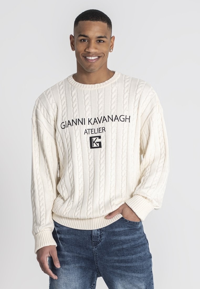 Cremefarbener, kabelstrick-gestrickter Pullover mit runder Ausschnittöffnung, gerippten Bündchen und gesticktem Logo, kombiniert mit blauen Jeans.