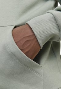 Sweatshirt vert clair avec des poignets côtelés. Une main est glissée dans une poche kangourou, mettant en valeur le tissu texturé et les détails de couture.