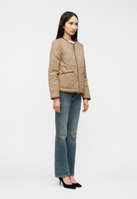 Μπεζ μπουφάν με καπιτονέ, στρογγυλή λαιμόκοψη, φερμουάρ μπροστά και δύο τσέπες. Συνδυασμένο με μπλε τζιν παντελόνι ίσιου ποδιού και μαύρες μυτερές γόβες.