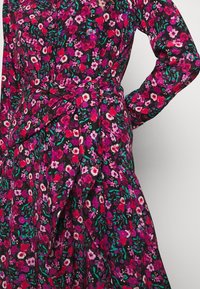 Schwarzes Kleid mit rotem, rosa und lila Blumenmuster, mit langen Ärmeln und einem breiten Stoffgürtel, der an der Taille gebunden ist.