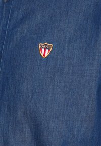 Camicia di denim blu scuro con finitura testurizzata. Presenta un logo GANT a forma di scudo, ricamato in oro, rosso e blu navy.