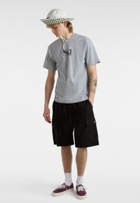 Grijs T-shirt met korte mouwen en een logografiek; zwarte cargoshorts; witte vissershoed met groen geblokt bandje; bordeauxrode sneakers met witte zolen.