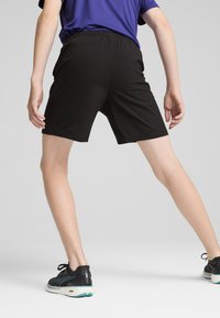 Svarta träningsshorts med elastisk midja, knä-lång längd och slät tyg; modell som bär svarta skor med turkosa accenter.