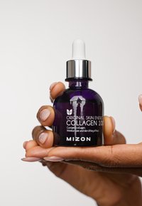 Lila glasdropperflaska märkt "Mizon Original Skin Energy Collagen 100", hålls i en hand, har ett silverfärgat lock och en svart bas.