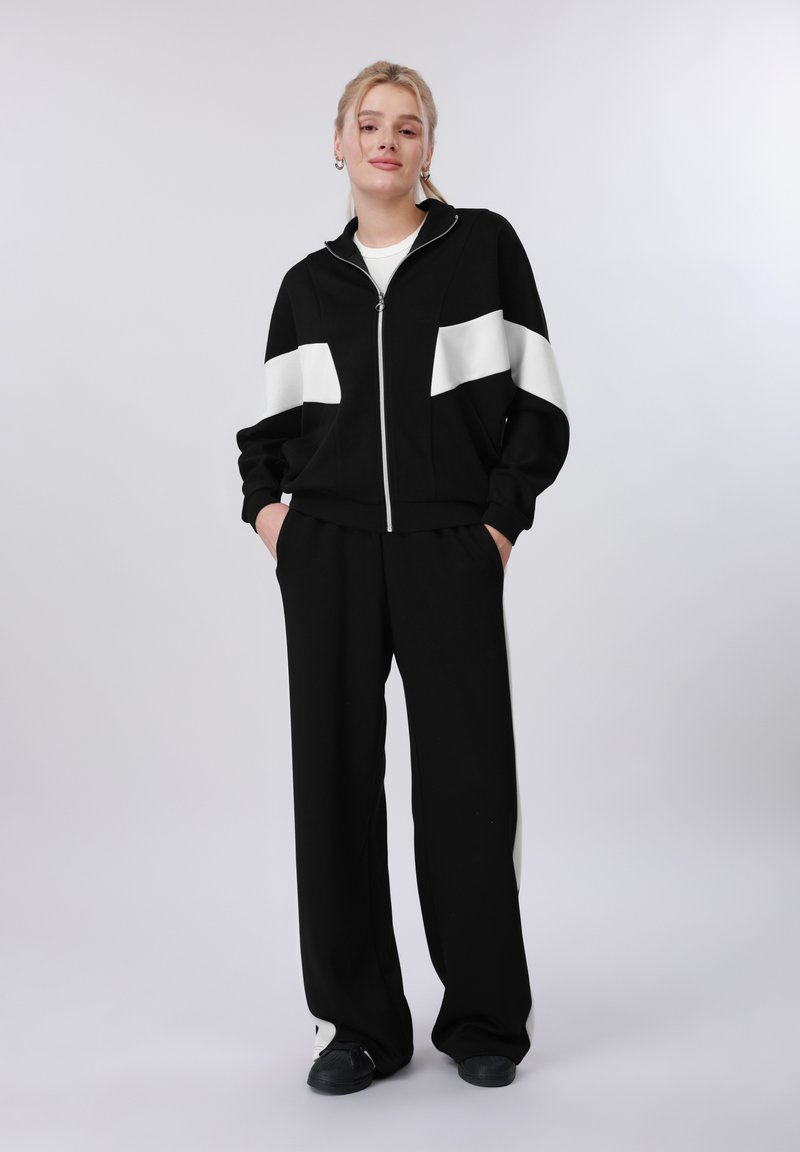 Jeune femme debout les mains dans les poches, portant une veste zippée noir et blanc, un pantalon large noir et des chaussures noires sur un fond uni.