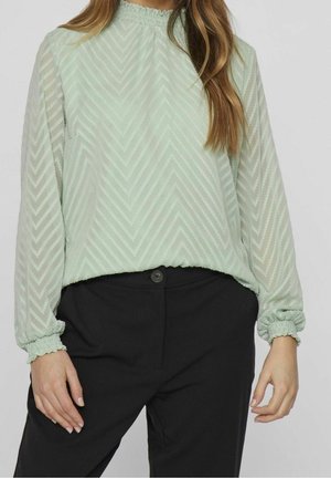 Femme portant un chemisier à manches longues vert clair à motif chevron avec des poignets élastiques, associé à un pantalon noir.