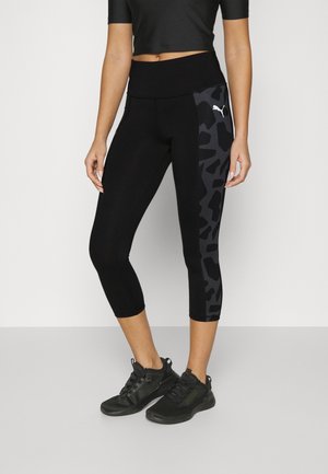 Femme portant des leggings courts noirs avec des panneaux latéraux sombres à motifs et des chaussures de sport noires, debout devant un fond uni.