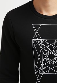 Chemise noire à manches longues présentant un motif géométrique de lignes blanches sur le devant. Encolure ronde côtelée et texture de tissu standard.