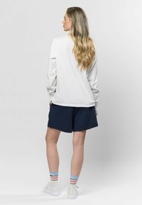 Biela košeľa s dlhým rukávom a námornícke šortky, so športovými bielymi topánkami a vzorovanými ponožkami s červenými, modrými a oranžovými pruhmi.